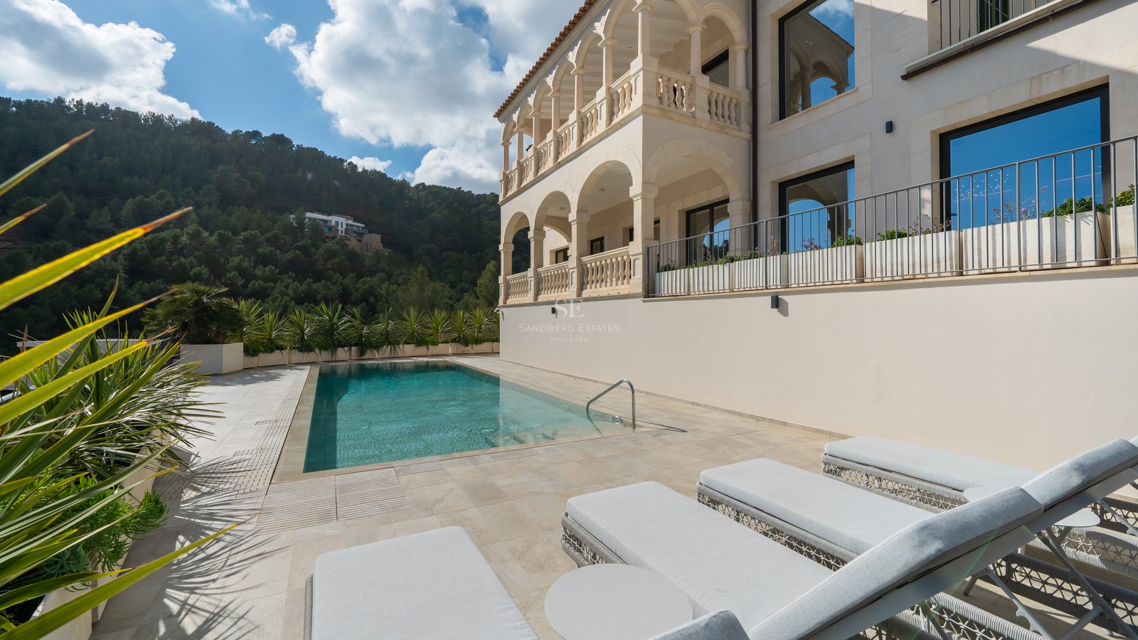 Villa de lujo con piscina rectangular, tumbonas, balcones de piedra con arcos y colinas verdes al fondo.