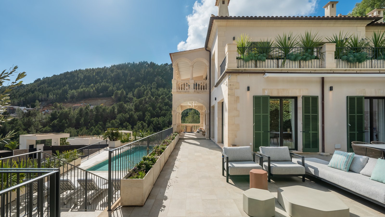 Terraza de lujo con mobiliario moderno, vistas a una piscina y montañas boscosas bajo un cielo despejado.