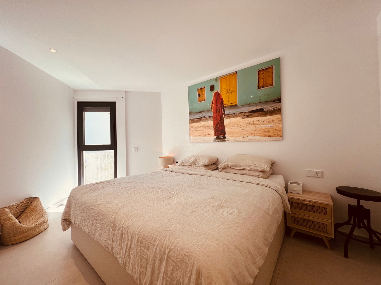 Minimalist bedroom featuring a large bed with beige linens, a wooden bedside table, and a large colorful wall painting.