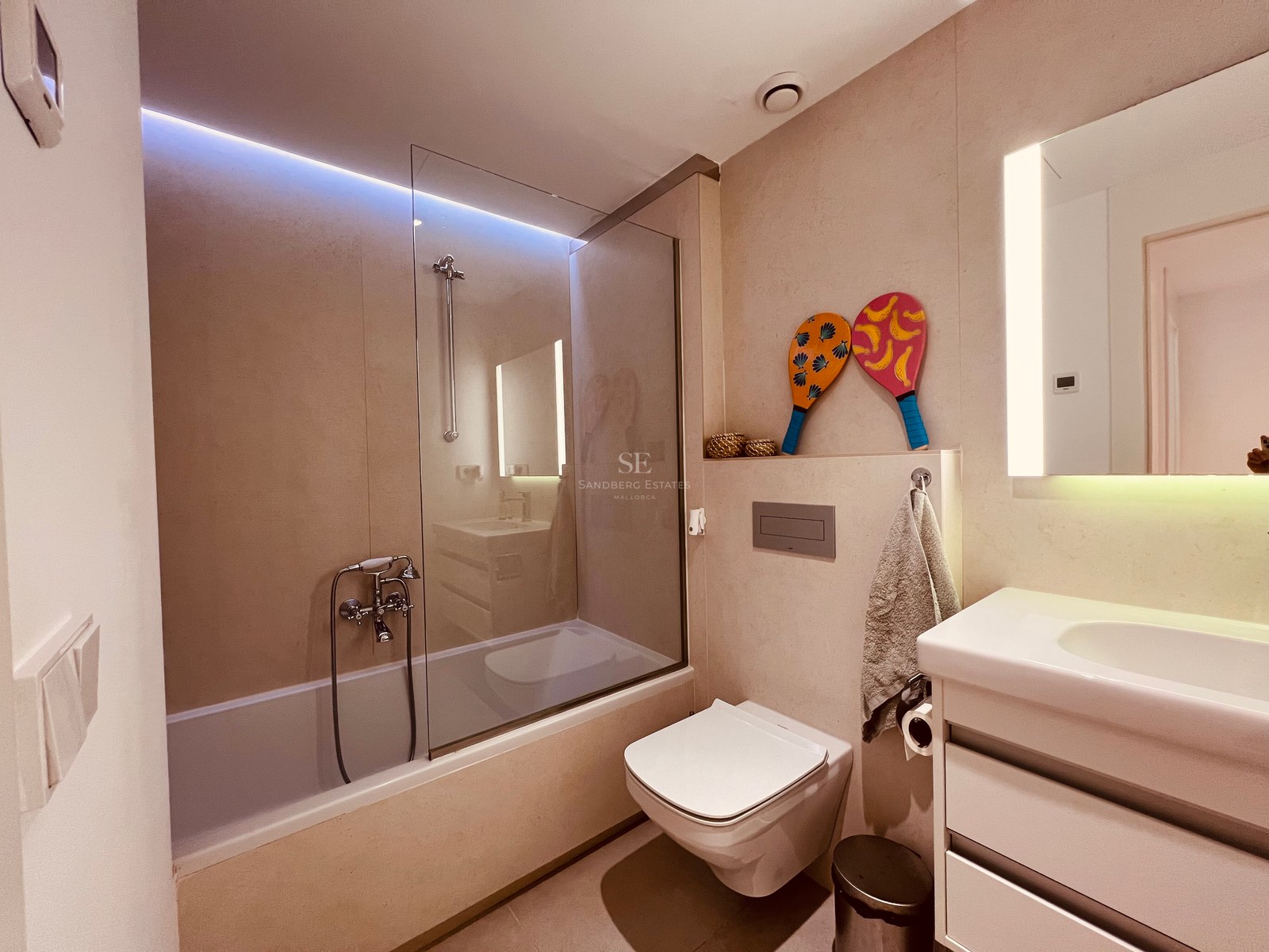 A contemporary bathroom featuring a bathtub with glass screen, floating toilet, and lighted vanity in neutral sand tones.