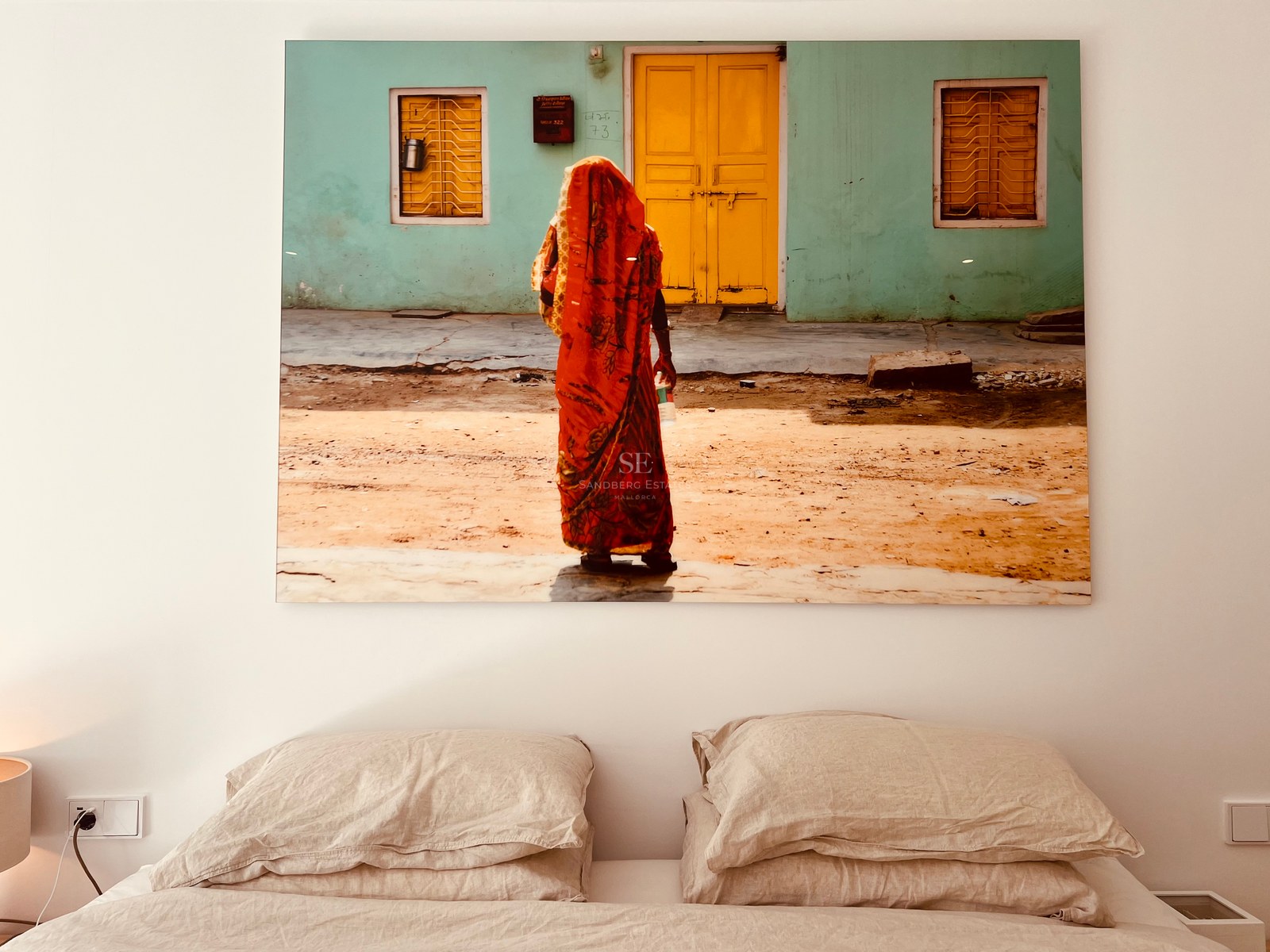 Large colorful artwork hanging above a bed with neutral linen pillows against a white wall.