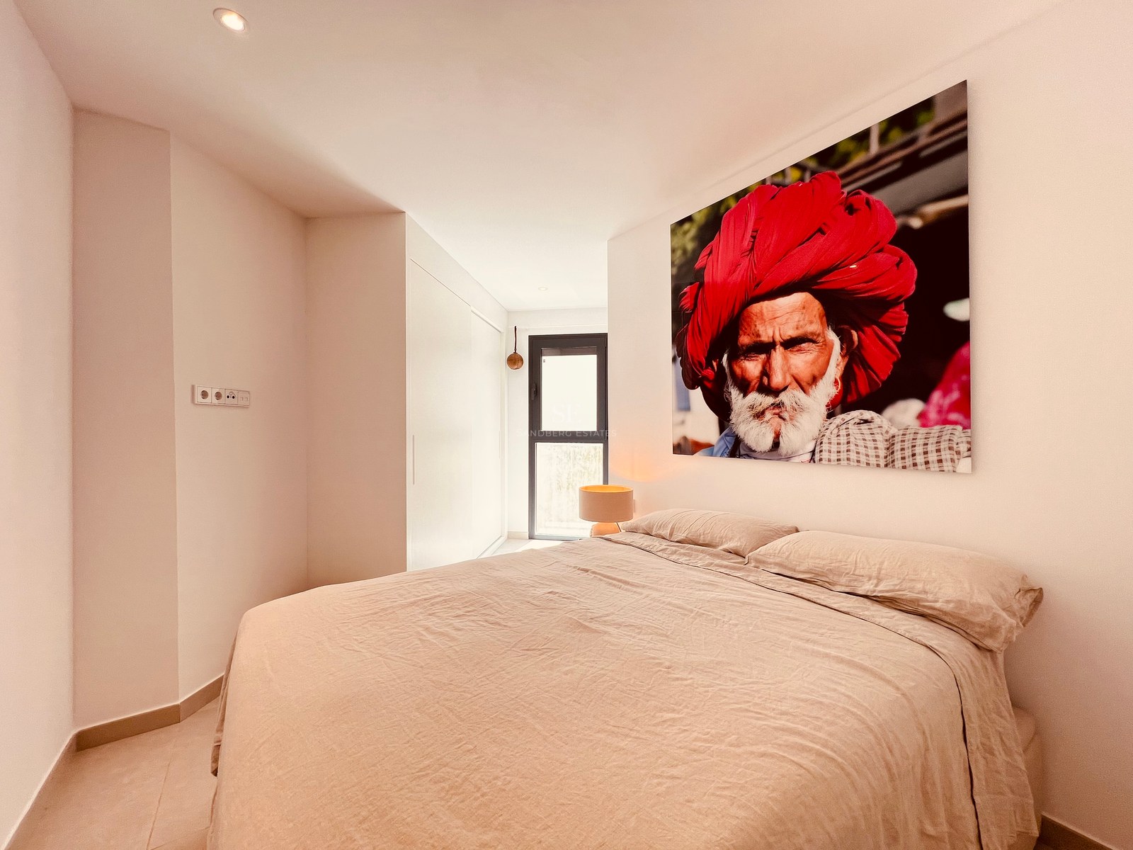 A modern bedroom featuring a large bed with beige linens and a vibrant portrait painting above the headboard.