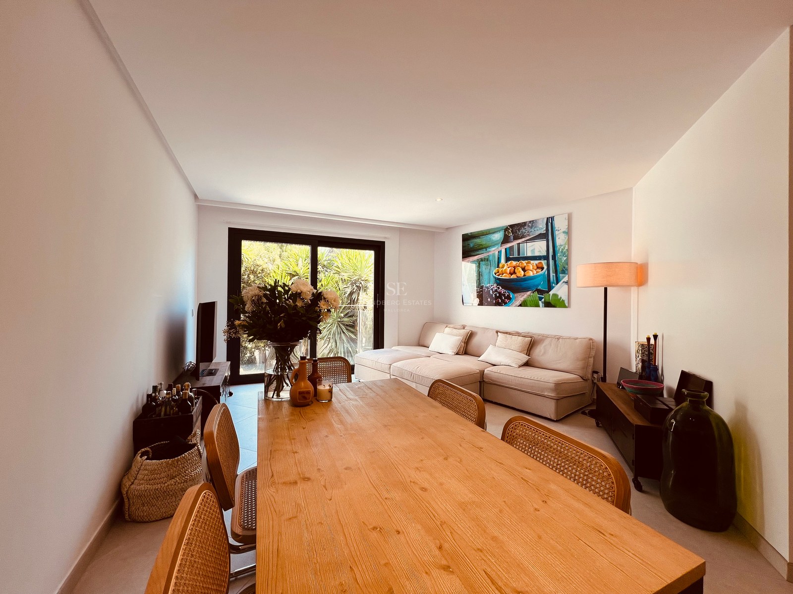 Living and dining area featuring a large wooden table, rattan chairs, and a beige sofa by sliding glass doors.