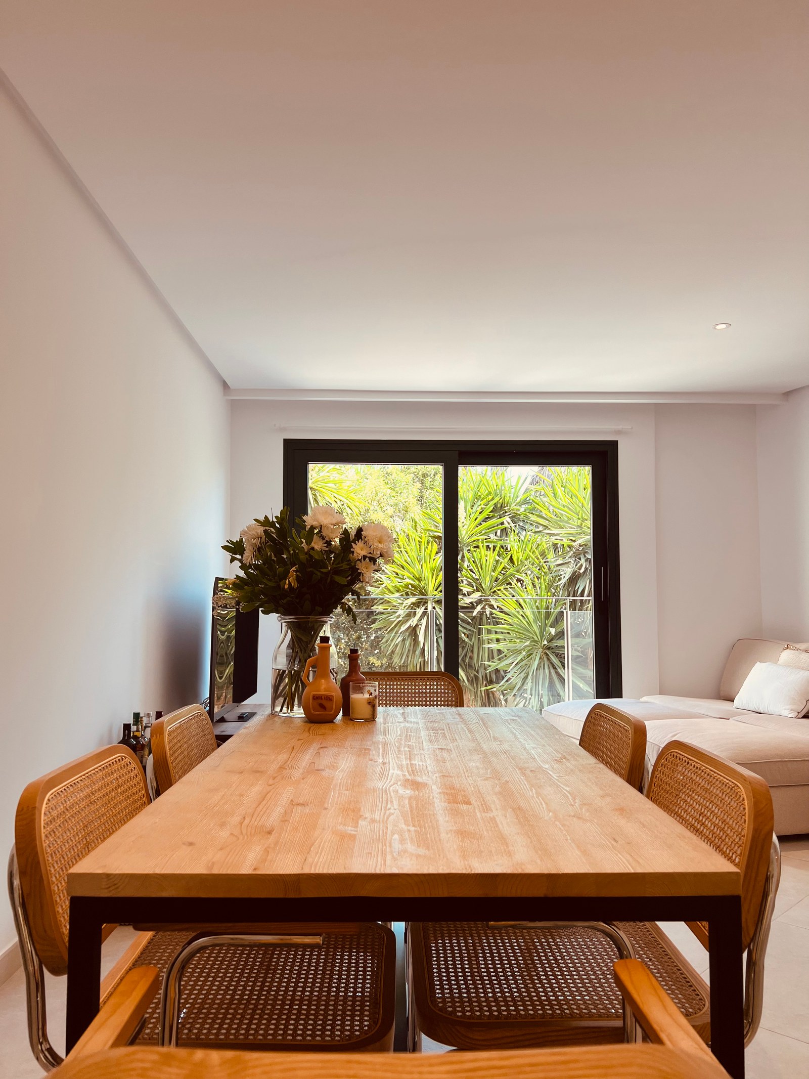 Light wood dining table with rattan chairs in front of a large window looking onto tropical greenery.