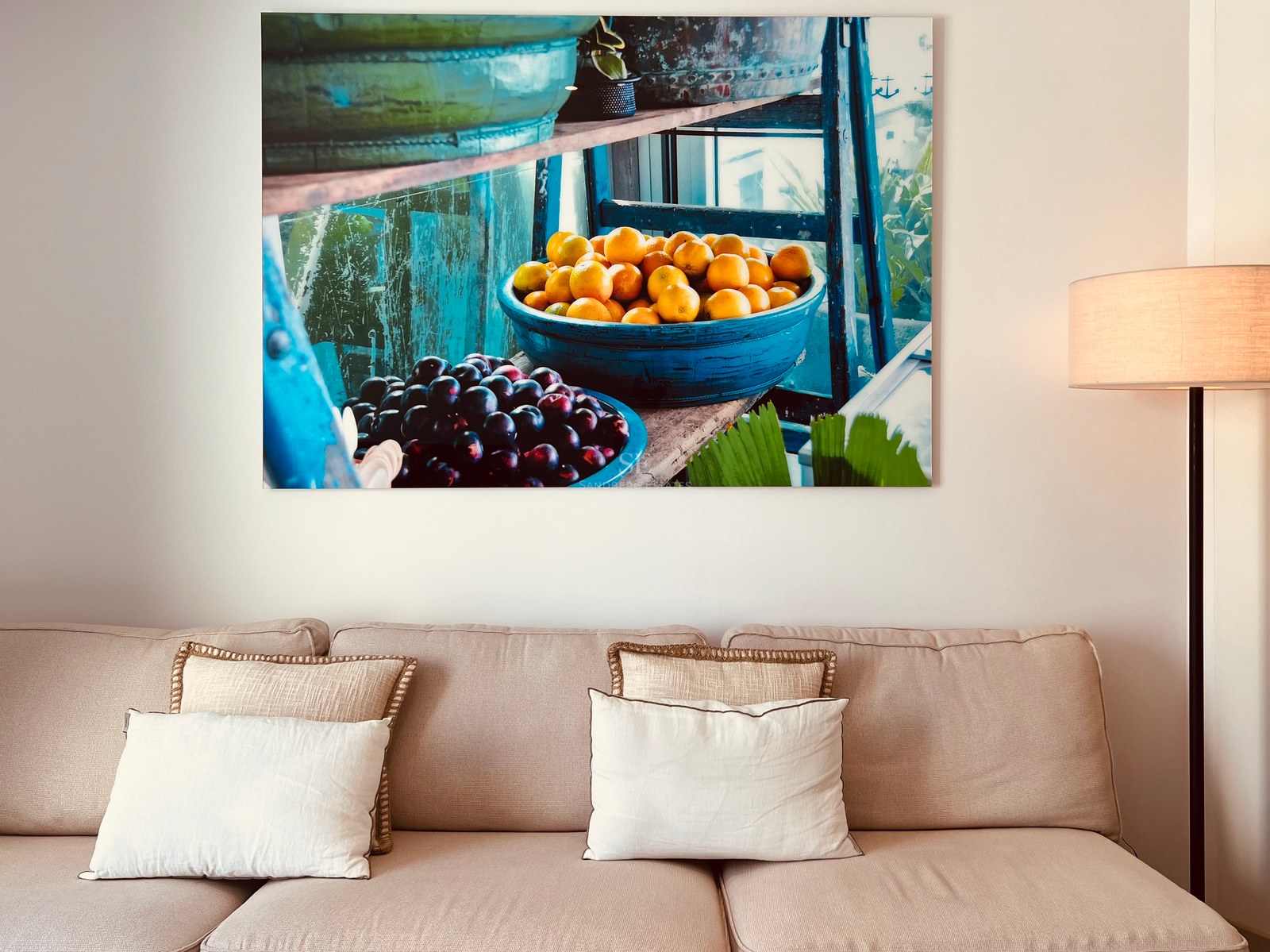 Beige sofa with decorative pillows below a large canvas painting of fruit in blue bowls.