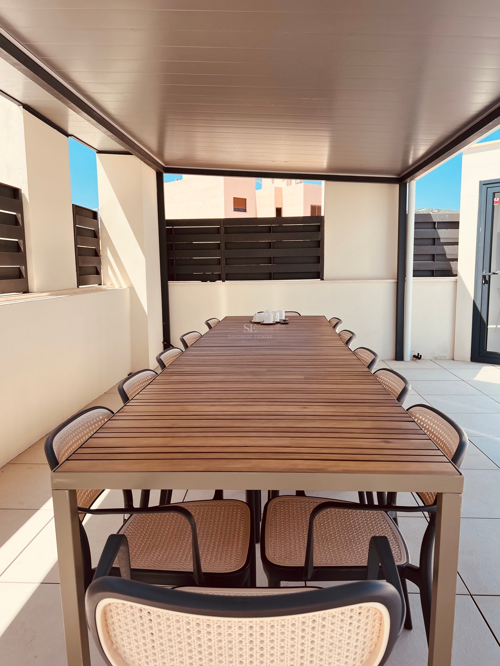 A long wooden outdoor dining table with ten chairs under a modern pergola on a sunlit terrace.