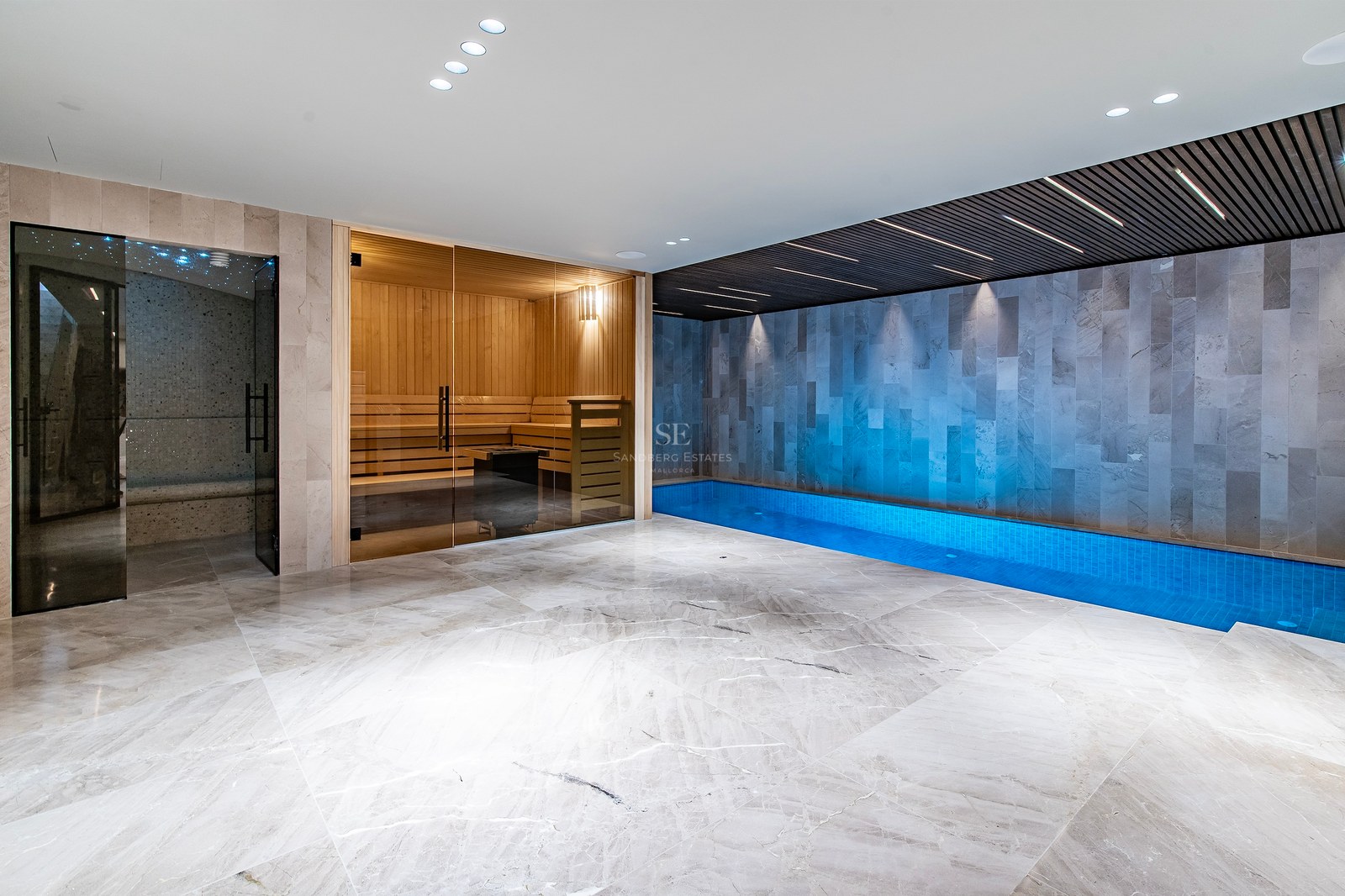 Modern indoor spa area featuring a marble floor, a glass-fronted sauna, and a long blue lap pool under accent lighting.