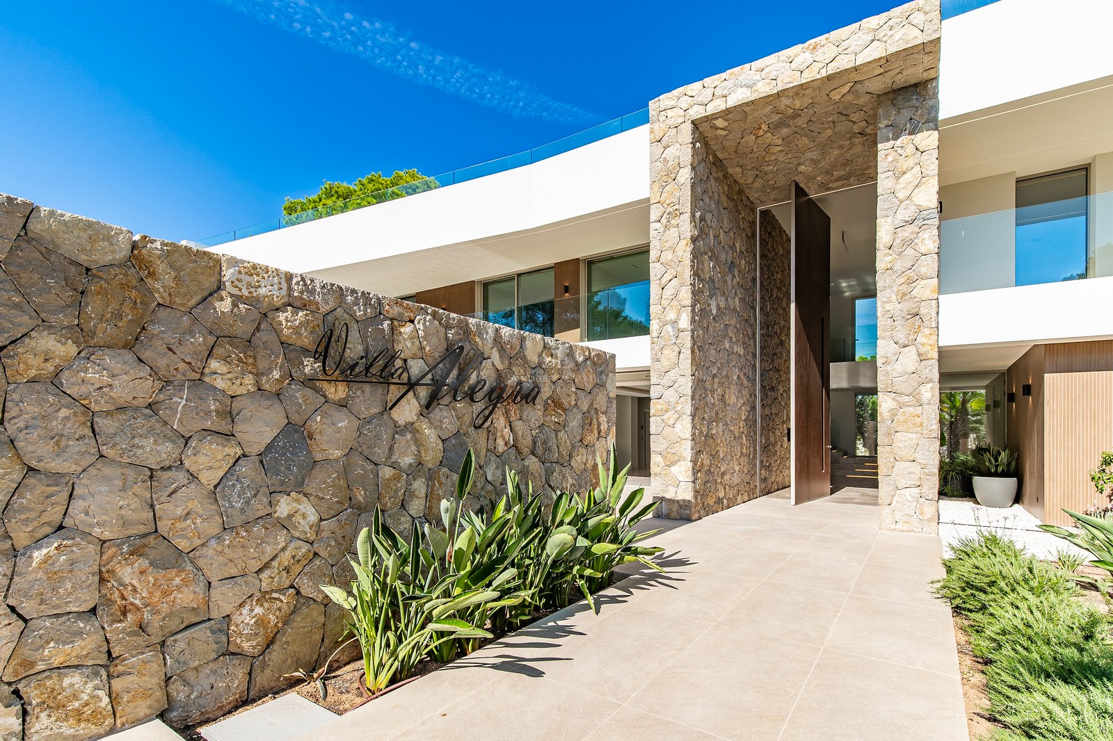 Modern villa entrance featuring a dry stone wall with Villa Alegra signage and a large wood pivot door.