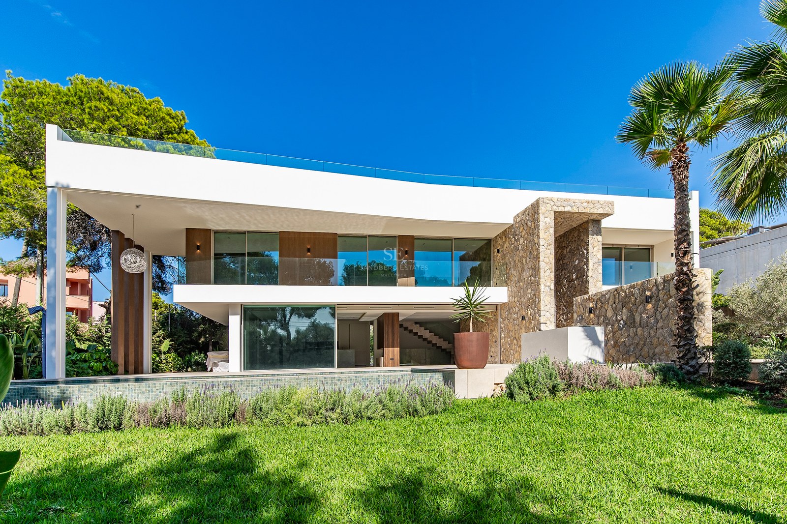 Modern white two-story villa with natural stone accents, glass balconies, and a swimming pool overlooking a lush green lawn.