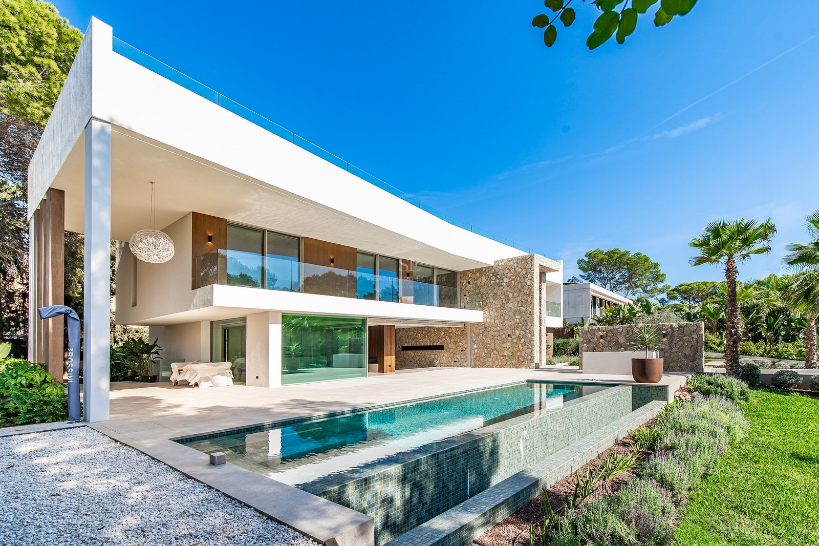 Modern two-story villa facade with infinity pool, natural stone walls, and large glass windows under a clear blue sky.