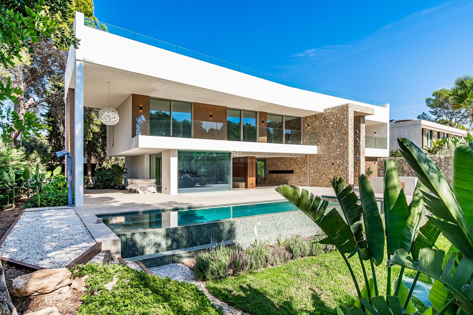 Two-story white modern villa with natural stone walls, a large swimming pool, and glass railings under a clear blue sky.