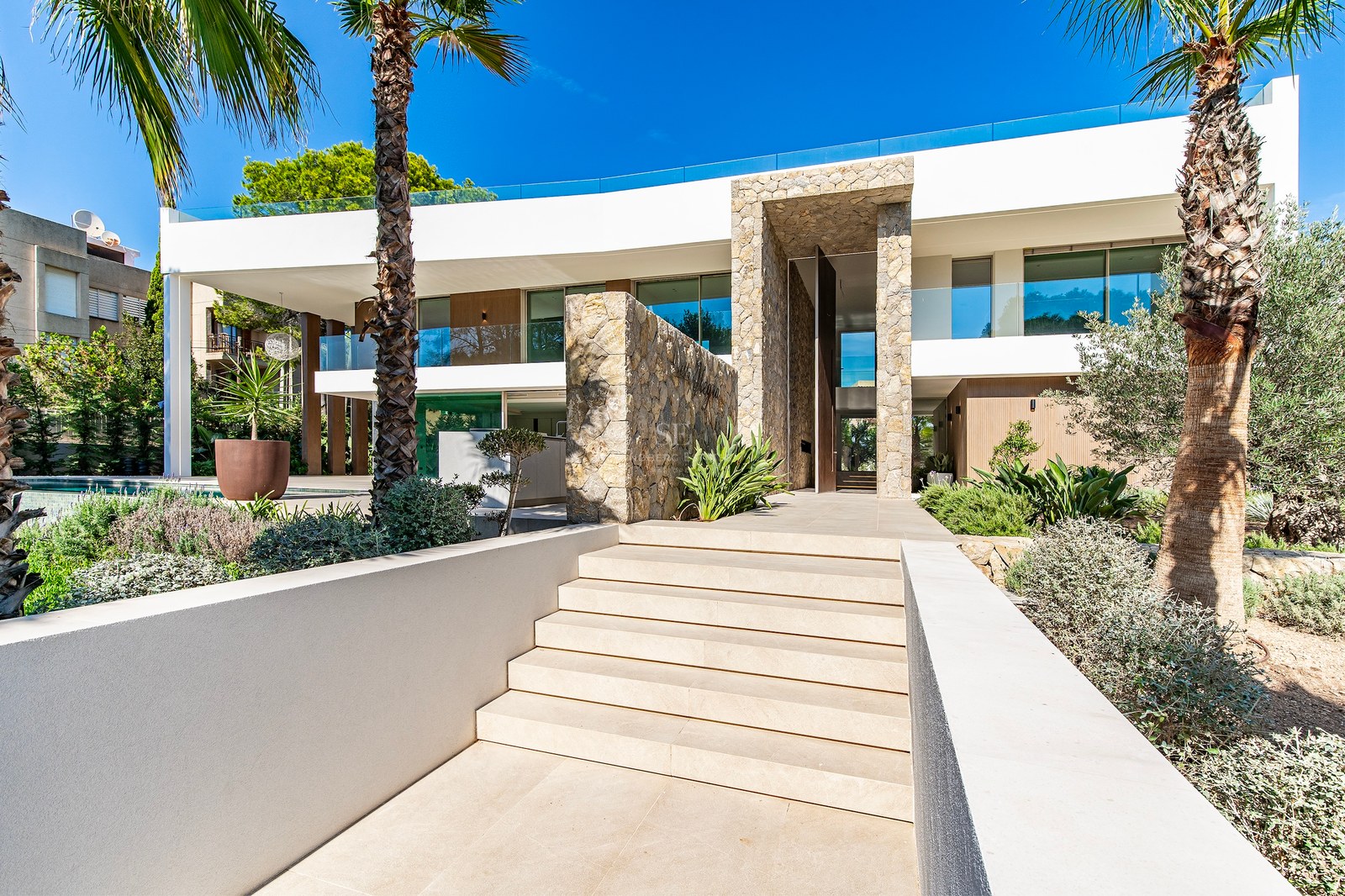 Modern white villa with natural stone feature walls, stone steps, and palm trees under a bright blue sky.