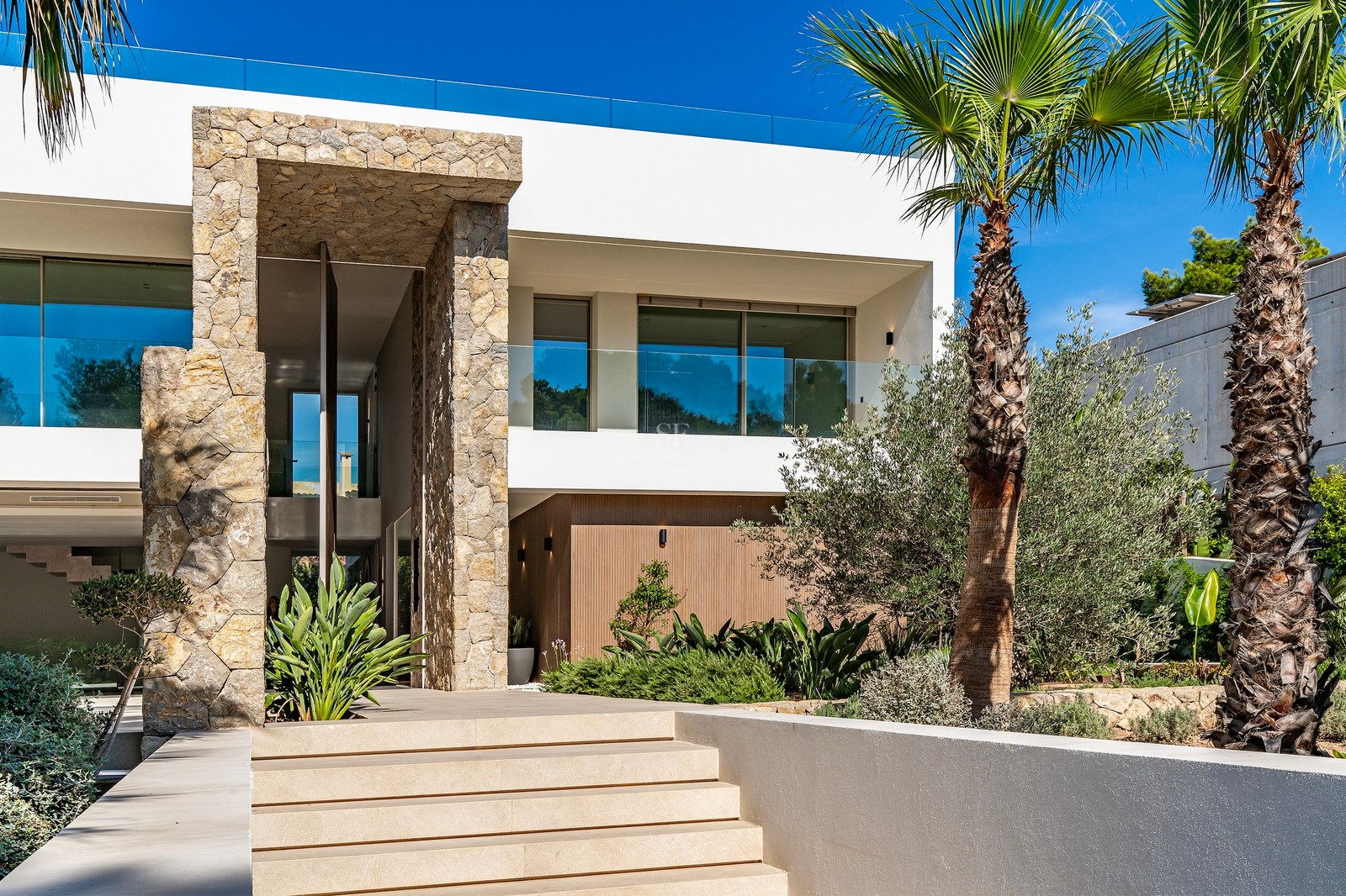 Modern villa entrance featuring wide stone steps, a high stone-clad portal, palm trees, and clear blue sky.
