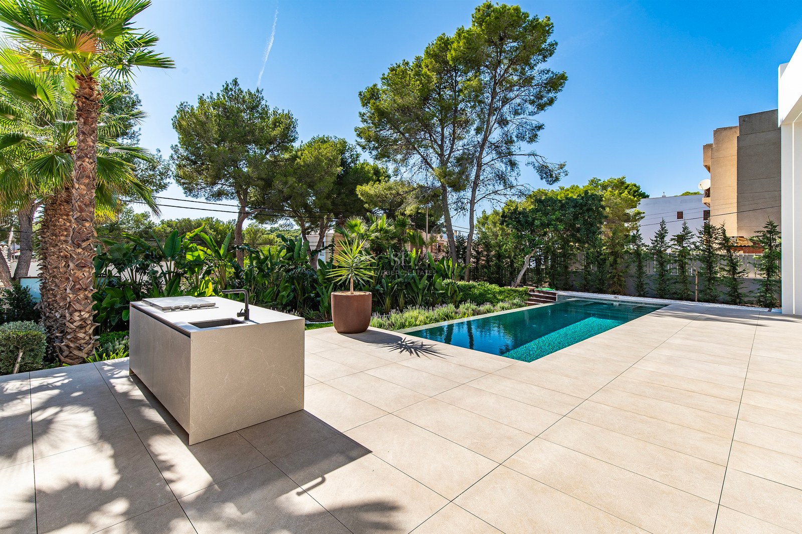 Stone terrace featuring a minimalist outdoor kitchen island next to a turquoise swimming pool and tropical gardens.