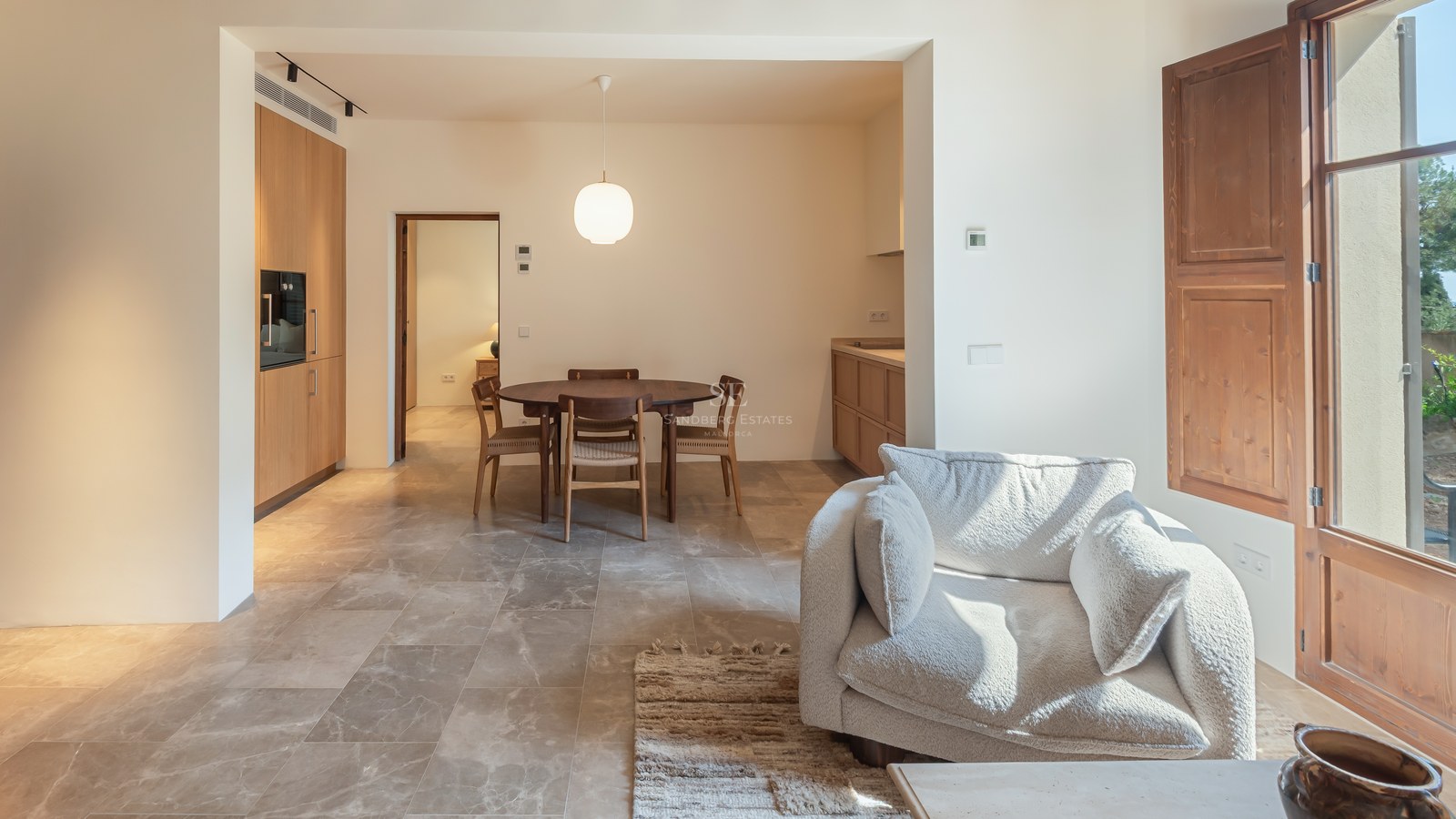 Salon moderne avec sol en pierre, menuiserie en bois, table ronde et fauteuil en bouclé blanc près d'une fenêtre.