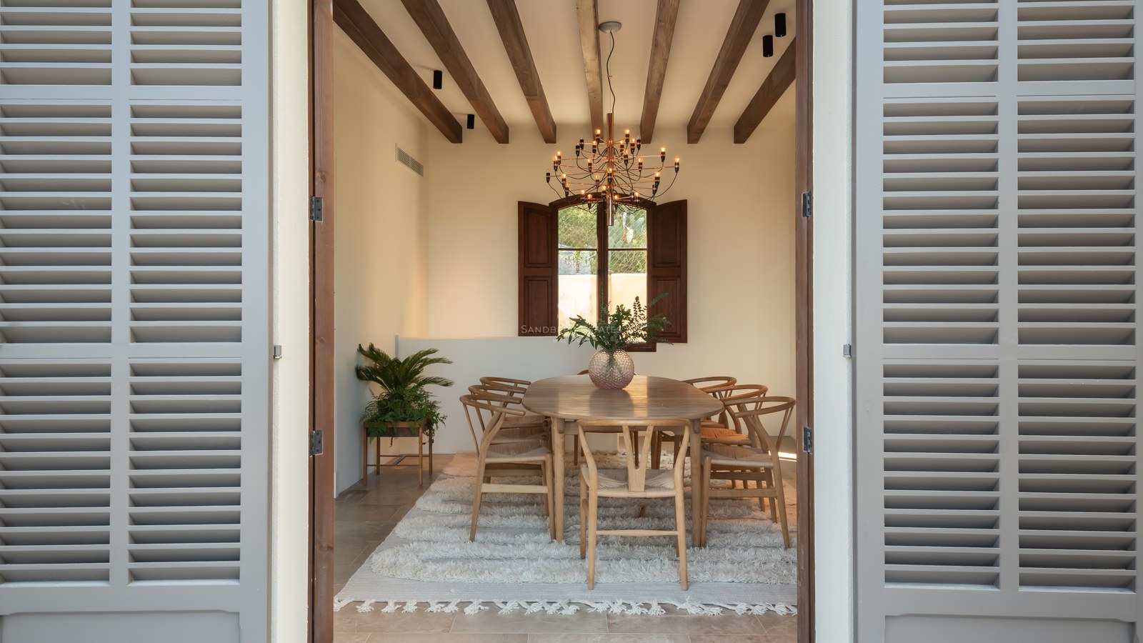 Salle à manger avec poutres apparentes, table en chêne, chaises Wishbone et tapis blanc encadrée par des volets gris.