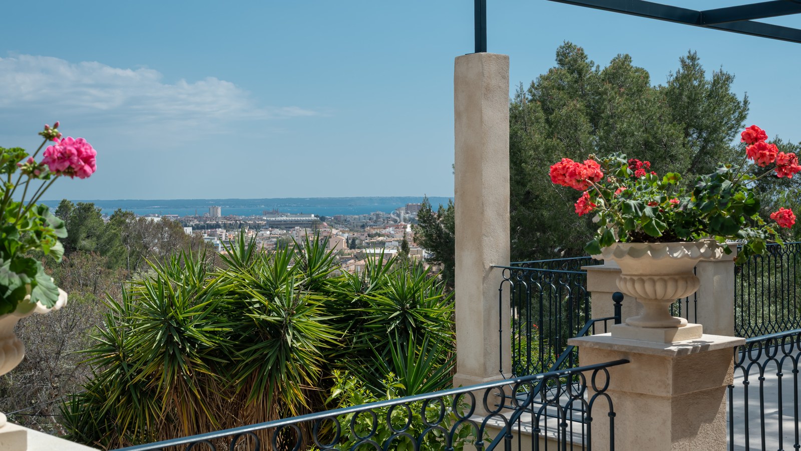 Vue depuis une terrasse sur une ville méditerranéenne et la mer, avec fleurs rouges et balustrade en fer forgé.