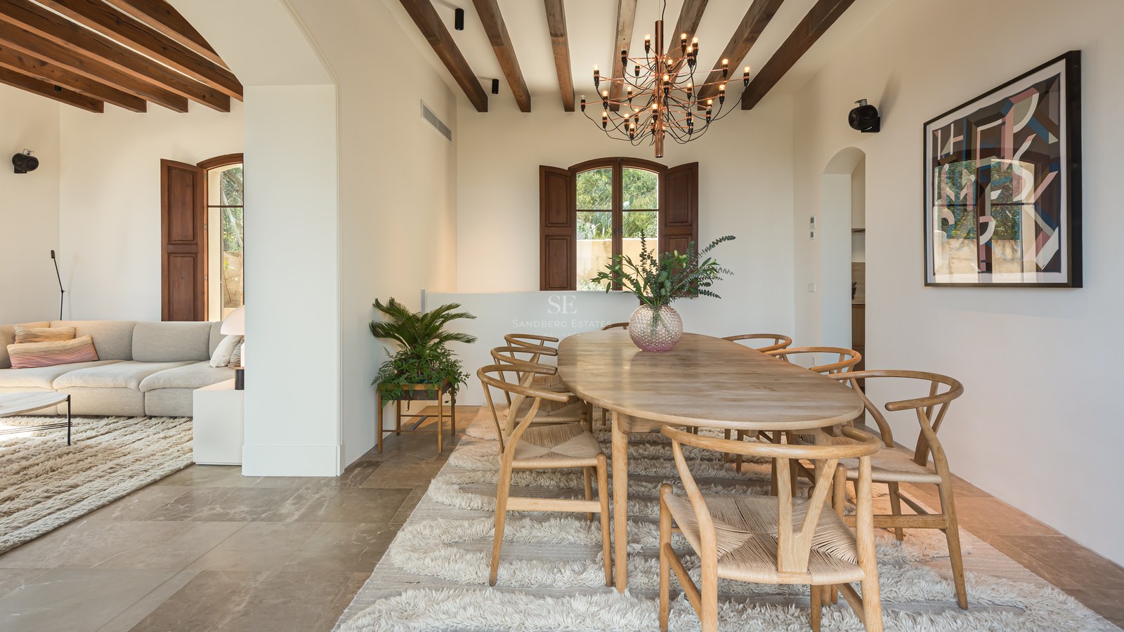 Salle à manger avec table ovale en bois, chaises style Wishbone, tapis en laine et plafond à poutres apparentes.