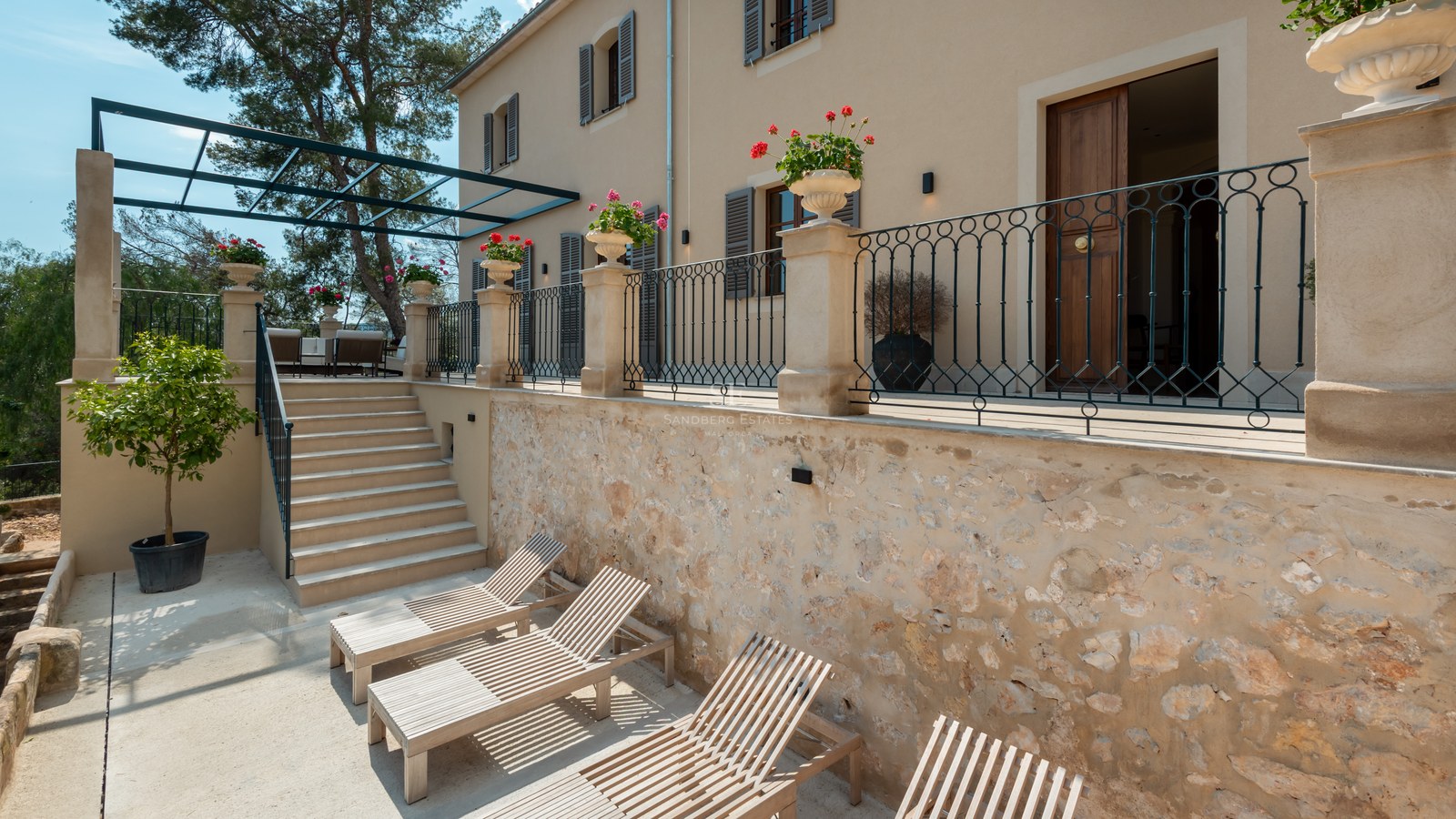 Terrasse en pierre à plusieurs niveaux avec chaises longues en bois, balustrades en fer forgé et façade de villa.