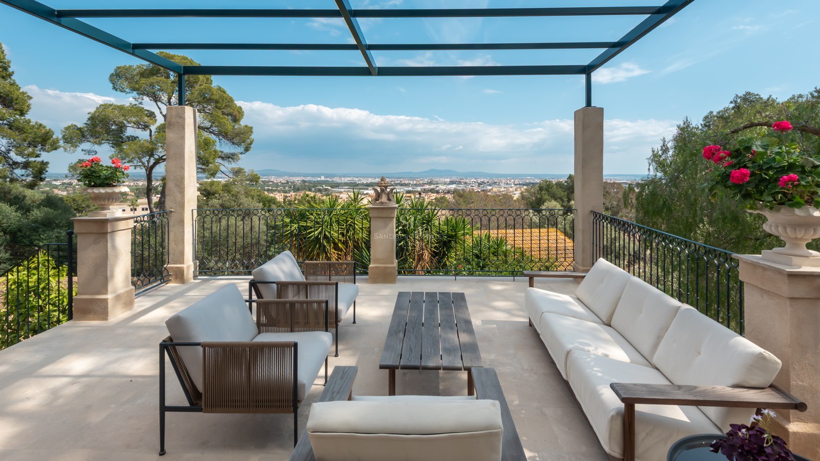 Terrasse de luxe avec canapés blancs, piliers en pierre et balustrade en fer forgé surplombant une vallée et des montagnes.