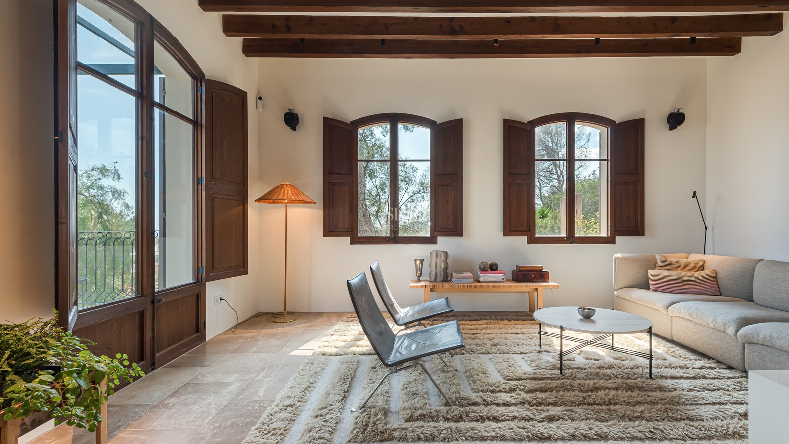 Salon lumineux avec sol en pierre, poutres en bois sombre, fenêtres cintrées avec volets et mobilier moderne.