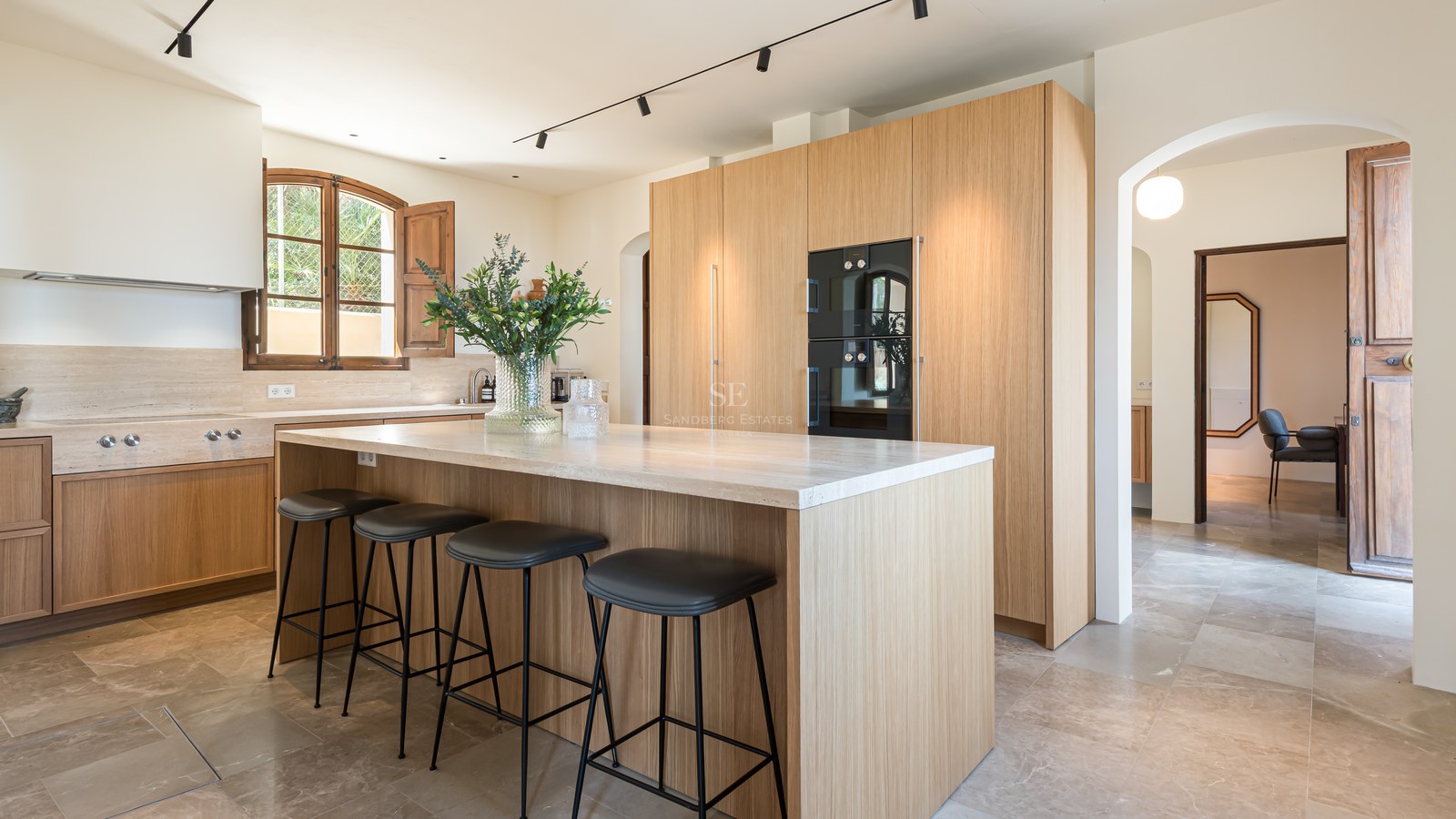 Cuisine contemporaine avec meubles en chêne, îlot en pierre naturelle, tabourets noirs et sol en marbre.