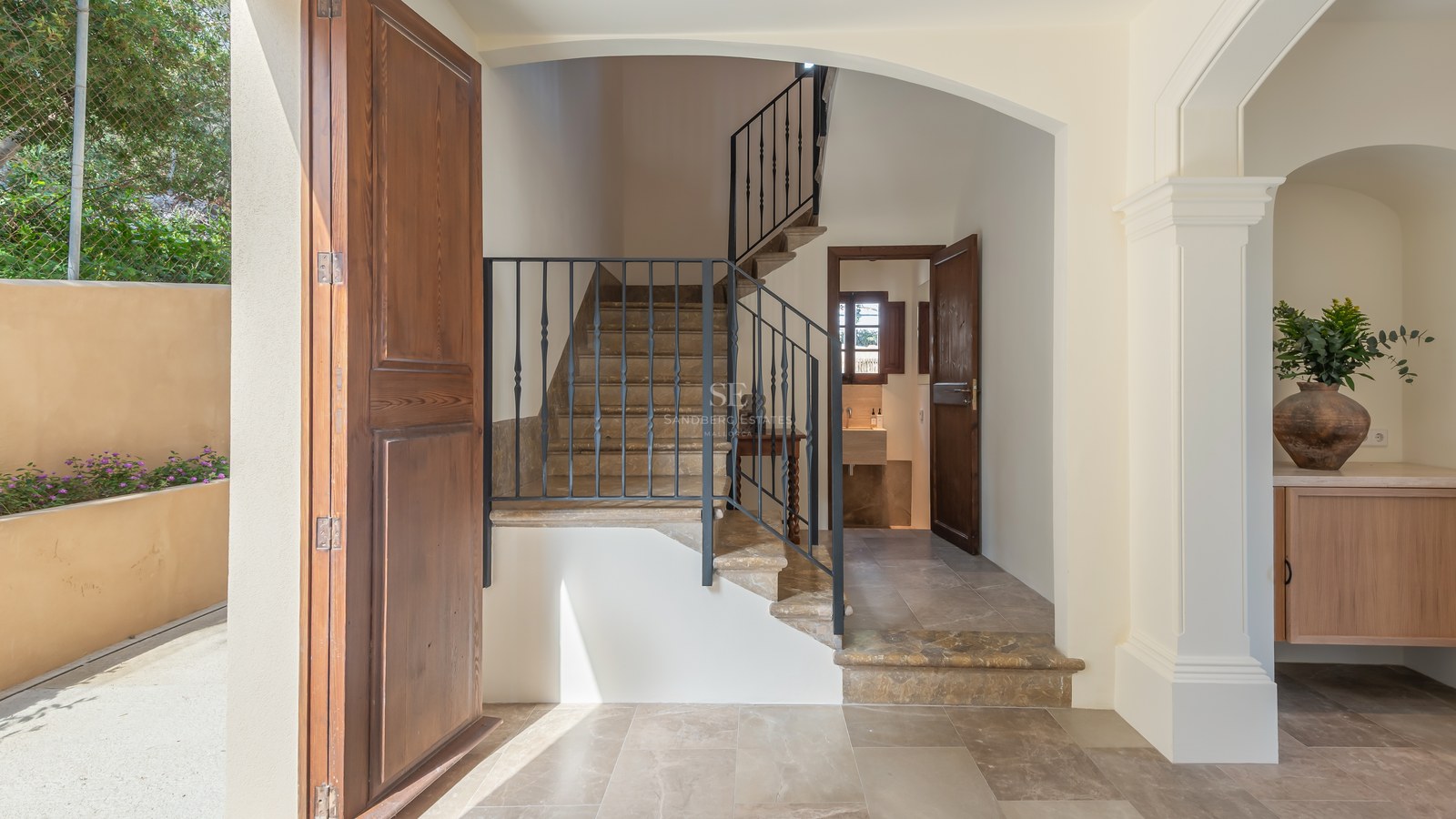 Entrée avec escalier en pierre, rampe en fer forgé, sols en marbre et porte en bois dans une villa de luxe.