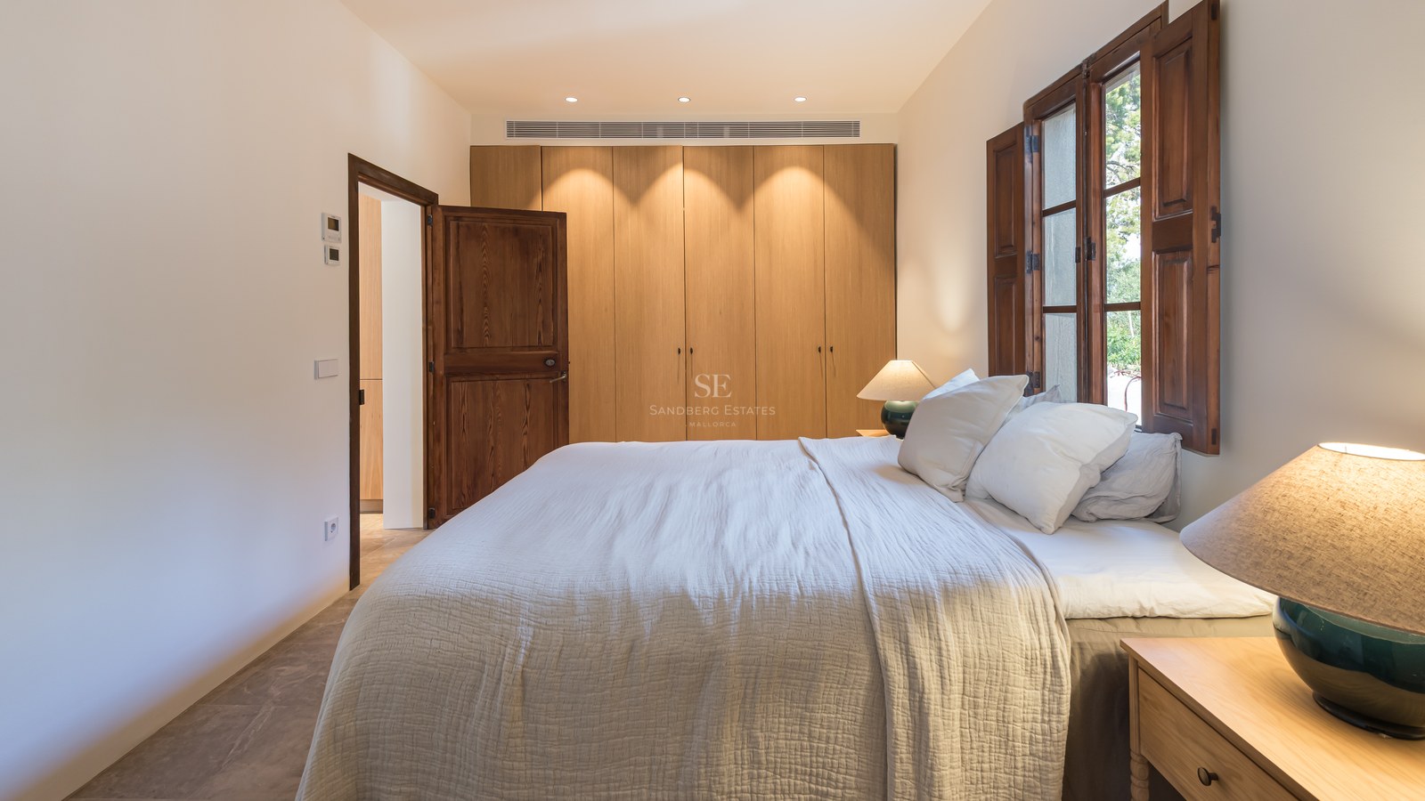 Chambre avec grand lit, placards intégrés en bois et volets majorquins sous une lumière chaleureuse.