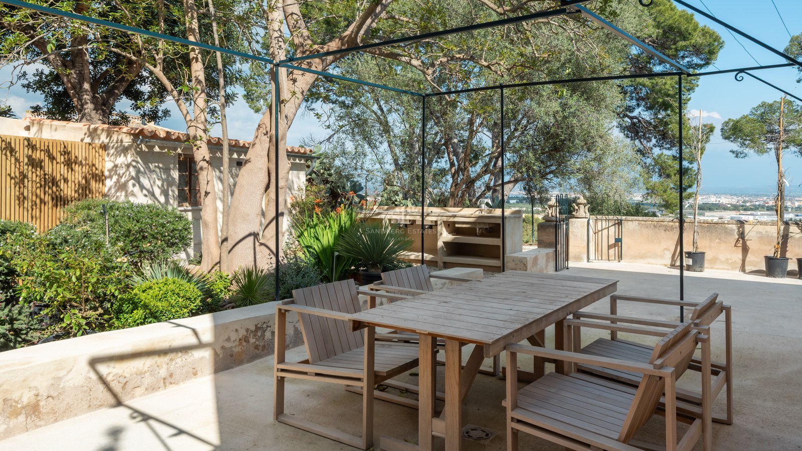 Espace dînatoire en bois sur terrasse en pierre avec pergola métallique, cuisine d'été et arbres matures.