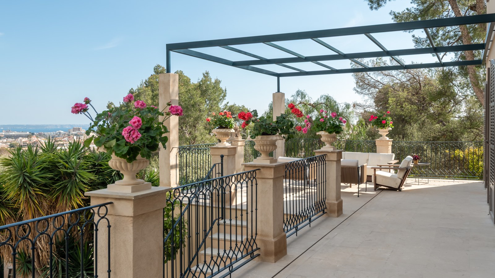 Terrasse en pierre avec garde-corps en fer, pergola métallique et fleurs surplombant la ville et la mer.