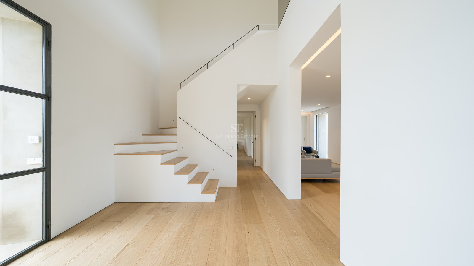 Ljus modern entréhall med naturligt ekparkettgolv och en minimalistisk vit trappa med smalt svart räcke.