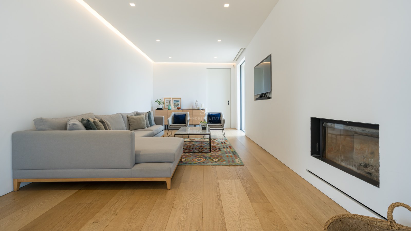 Ljust vardagsrum med ekparkett, grå L-sofa, inbyggd öppen spis och infälld LED-belysning.