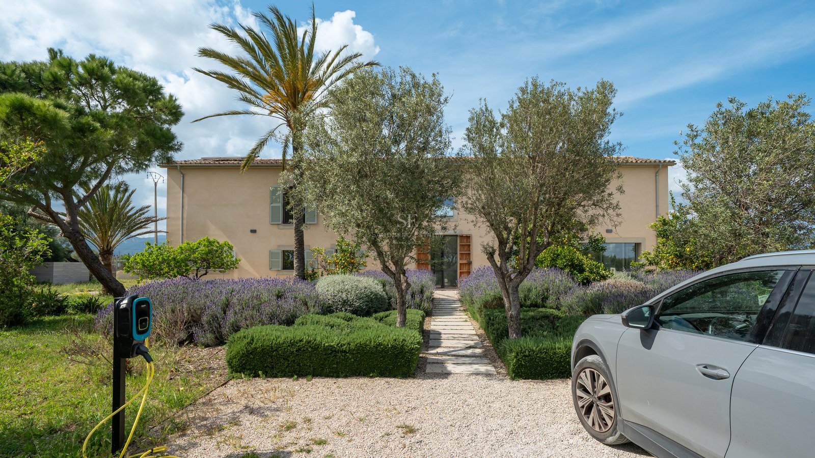 Fasad av sandfärgad villa med lavendelträdgård, stenstig och elbilsladdare på uppfarten.