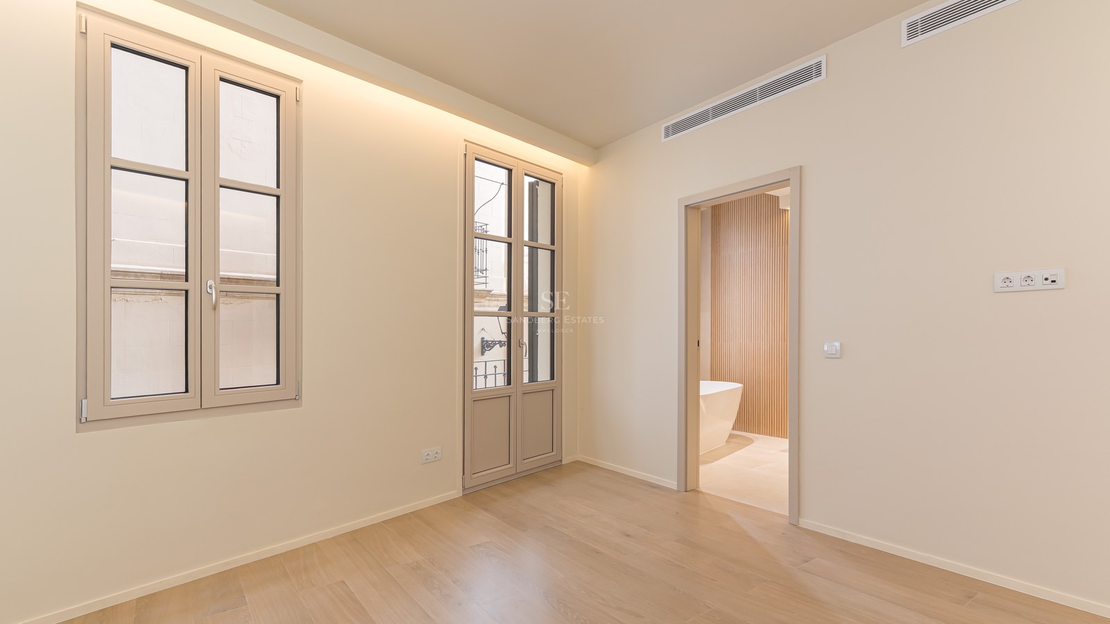 Dormitorio minimalista con suelos de madera clara, ventanales altos y vista a un baño con bañera exenta y listones de madera.