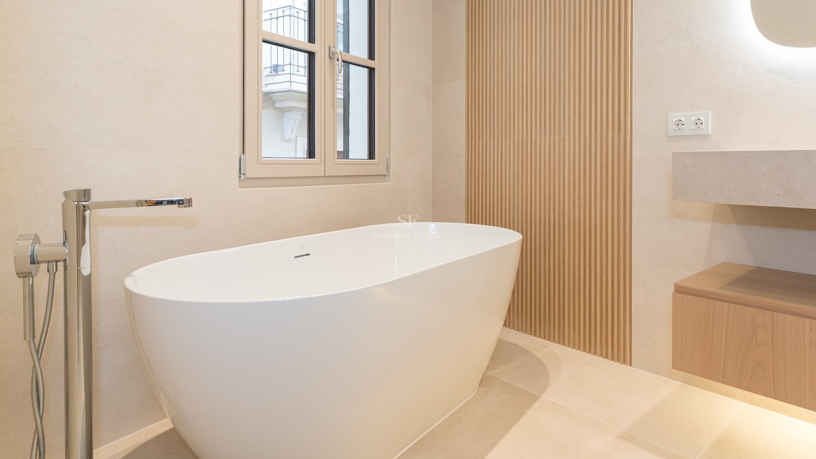 Bañera blanca exenta junto a pared de listones de madera y grifería cromada sobre suelo de piedra natural.