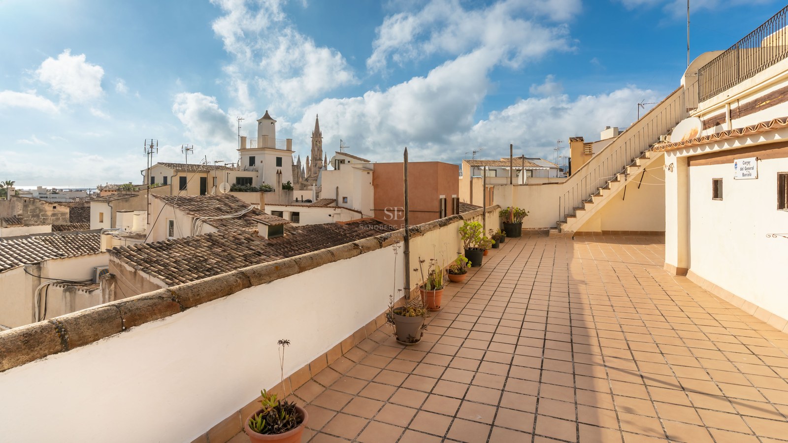 Terraza en azotea con baldosas de terracota con vistas a edificios históricos y un campanario gótico bajo un cielo azul.