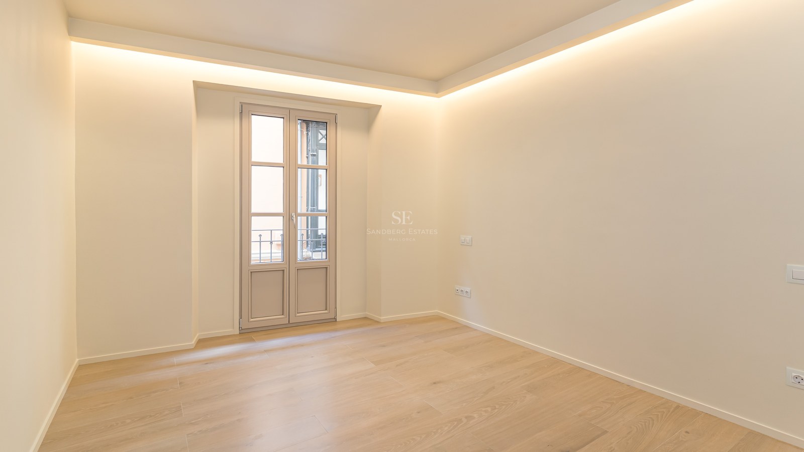 Habitación moderna vacía con suelos de roble claro, paredes crema, iluminación LED perimetral y balcón francés.
