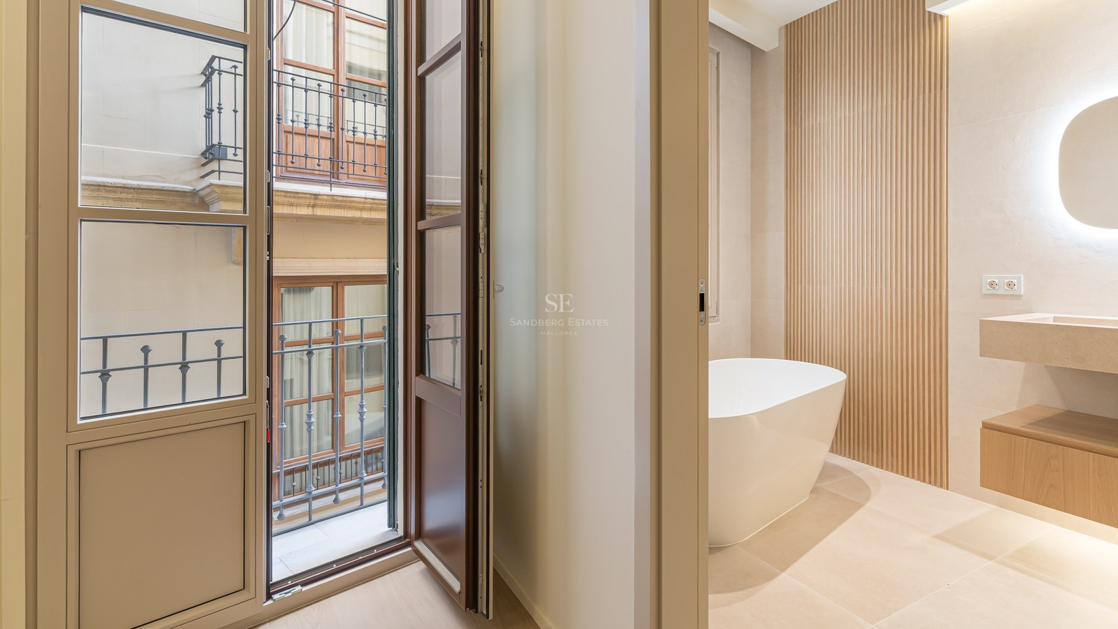 Baño moderno con bañera blanca exenta, lavabo de piedra, pared de listones de madera y balcón hacia patio exterior.