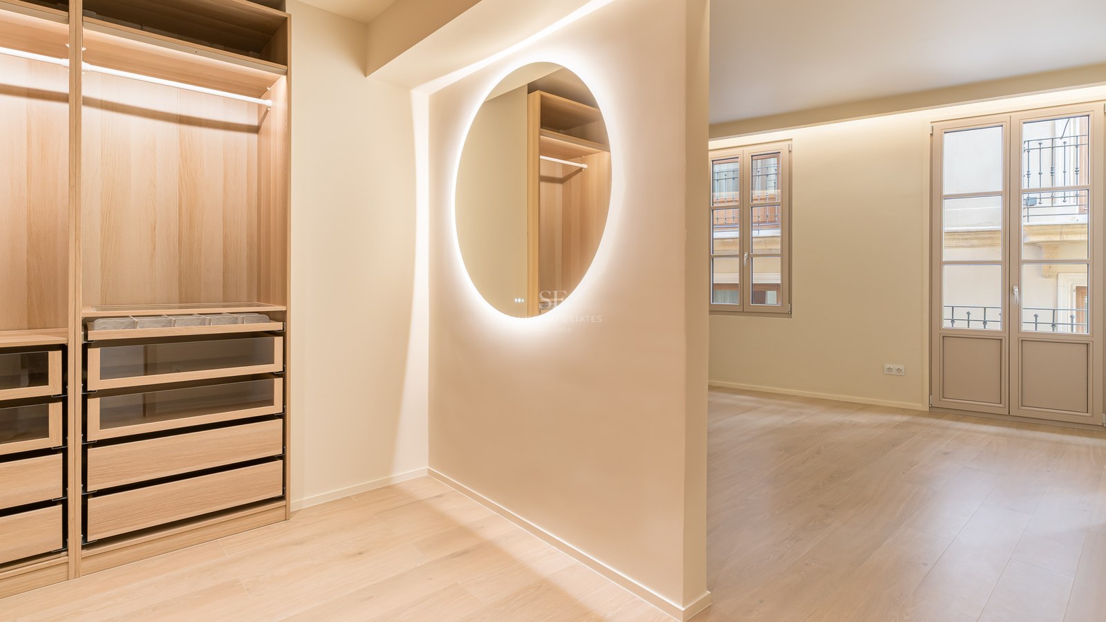 Vestidor de lujo con armarios de roble claro, espejo circular retroiluminado y cajones con frente de cristal.