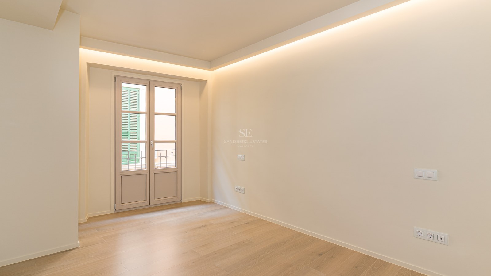 Dormitorio vacío con suelo de roble claro, paredes crema, iluminación indirecta y puerta hacia balcón francés.