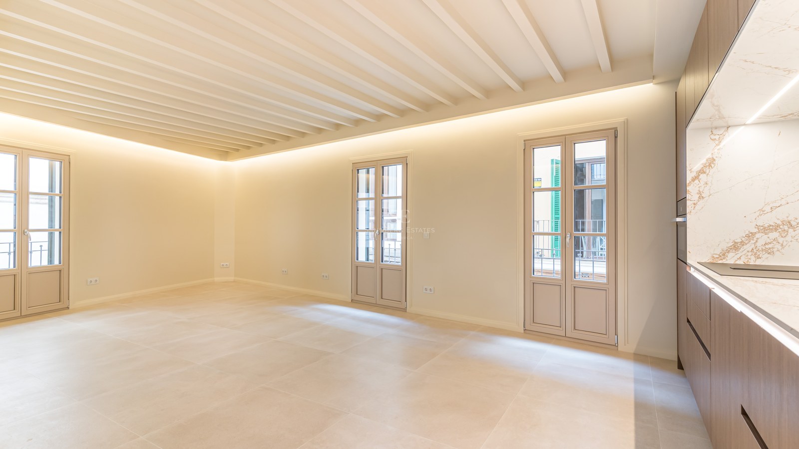 Salón vacío y luminoso con suelo de gres crema, techo de vigas vistas, tres puertas balconeras y cocina moderna lateral.