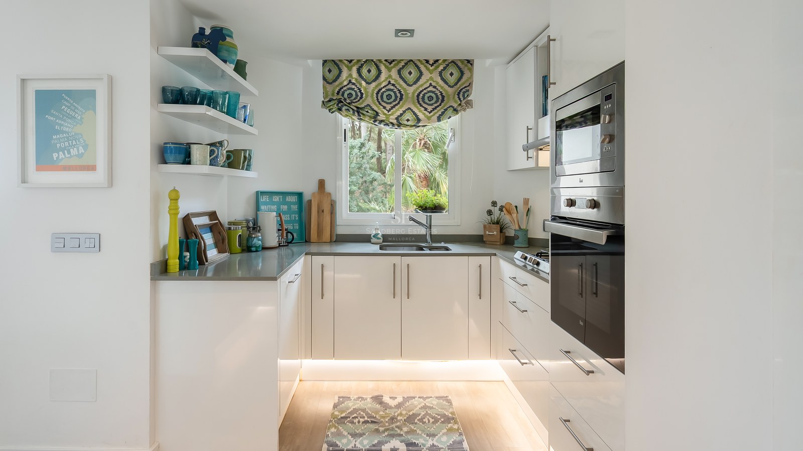Bright U-shaped kitchen with white cabinets, stainless steel appliances, and a window overlooking lush green trees.