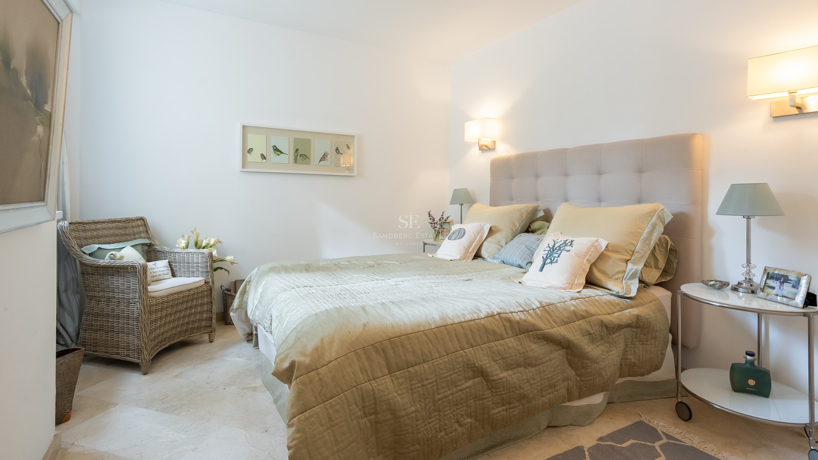A bright bedroom featuring a tufted headboard, sage green bedding, marble floors, and a wicker armchair.