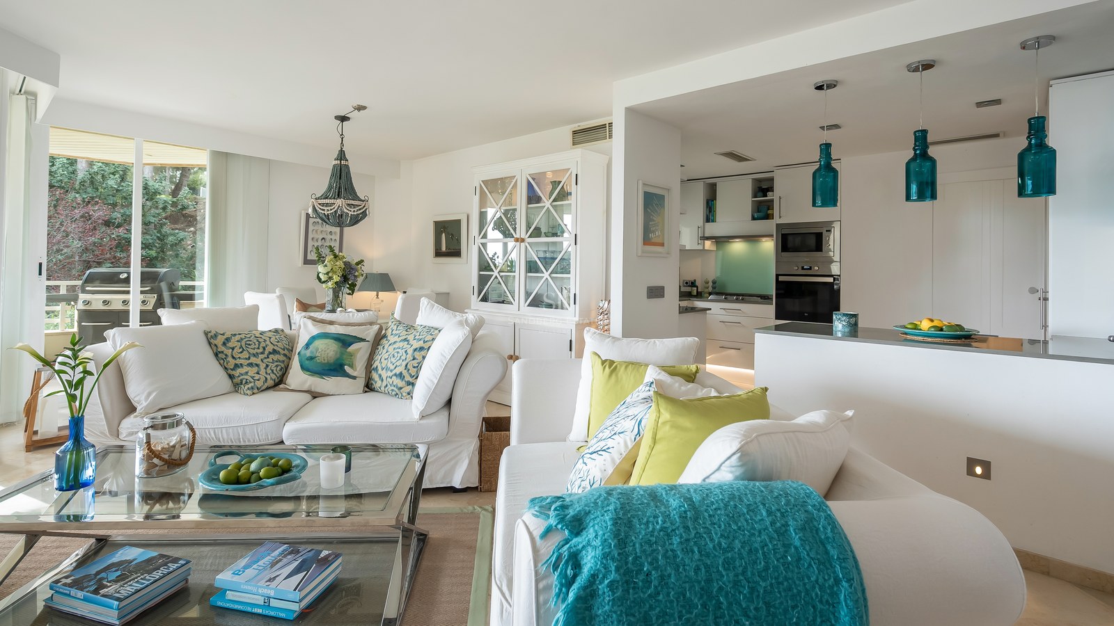 A sun-drenched living room with white sofas, turquoise accents, and a modern kitchen with blue pendant lights.
