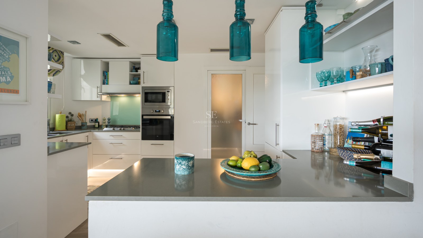 Modern white kitchen with grey countertops, turquoise glass pendant lights, and integrated appliances.