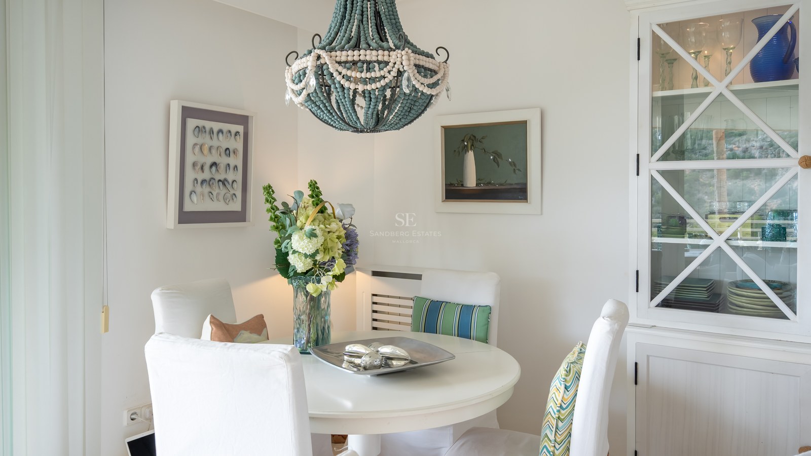 White round table, slipcovered chairs, blue beaded chandelier, and built-in cabinet in a bright dining area.