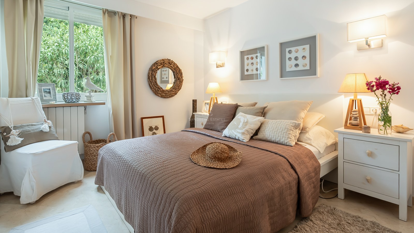 Bright bedroom with brown quilted bed, white nightstands, woven wall mirror, and garden views through a large window.
