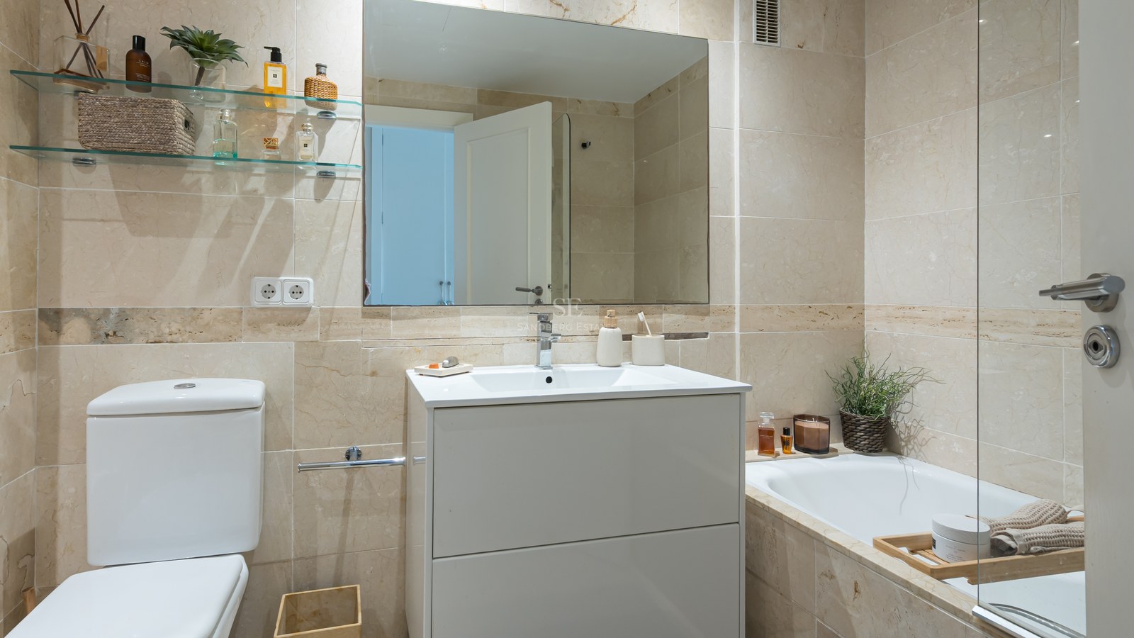 Bright bathroom featuring cream marble wall tiles, a white vanity unit, large mirror, and a bathtub with glass partition.
