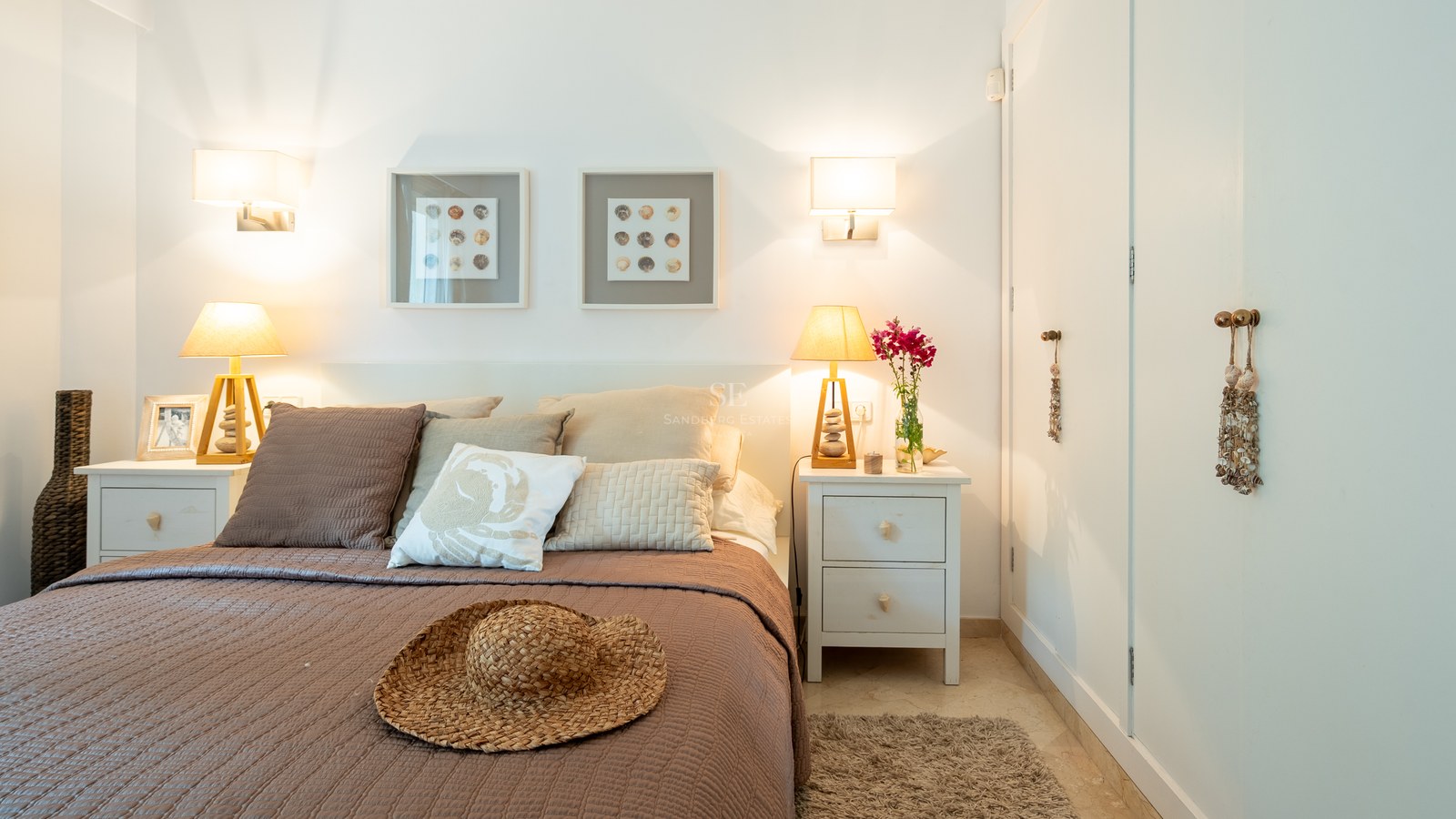 Bright bedroom featuring a large bed, wooden lamps, built-in white wardrobes, and coastal-themed decor.