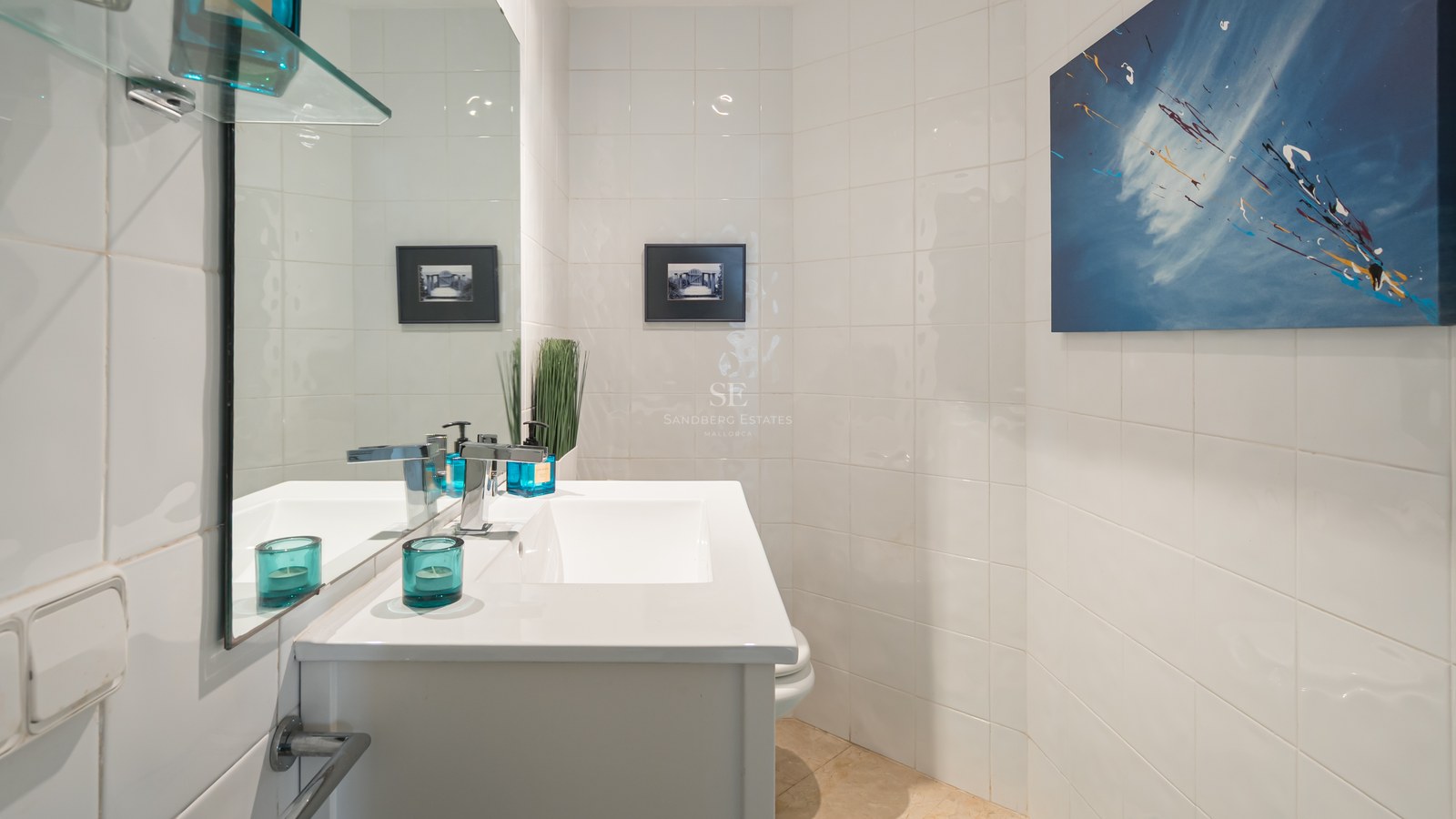 White tiled bathroom with a white vanity sink, chrome faucet, large mirror, and blue abstract wall art.